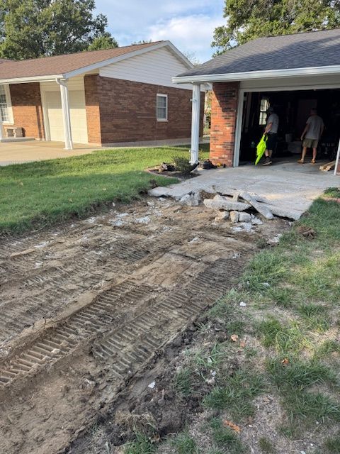 Dirt driveway with tire tracks, partial concrete removal next to garage; two people inside.