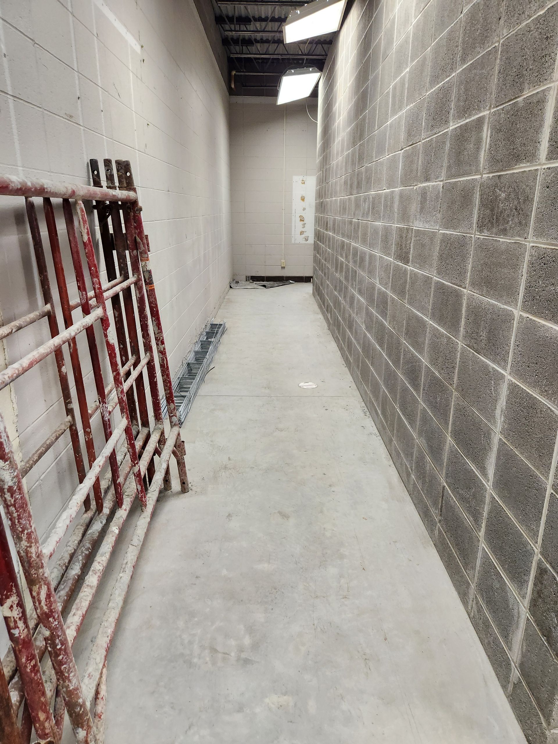 Narrow, concrete-floored hallway with cinder block and plain walls. Red scaffolding leans against one wall.