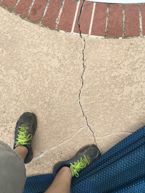 Person's feet with green-laced shoes stand near a long crack in a tan concrete surface next to a brick border.