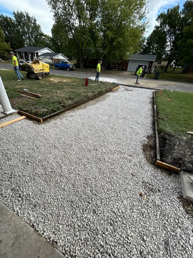 Gravel walkway construction. Workers in safety vests are laying gravel, with wooden borders defining the path.