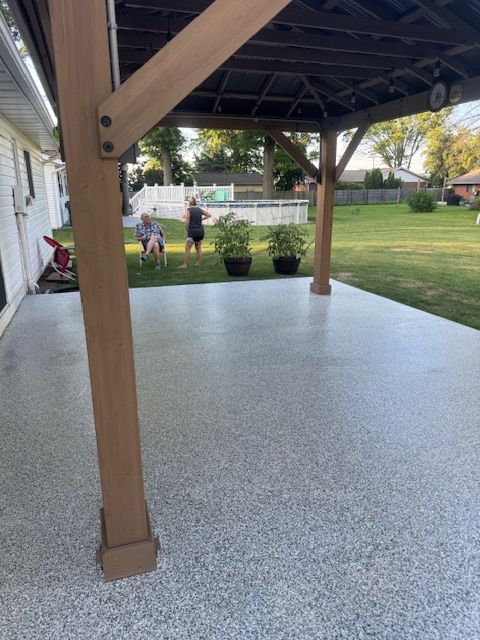 Concrete patio with flecks under a wooden gazebo, two people in the yard.