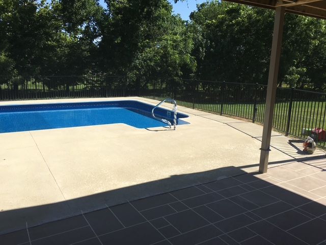 Swimming pool with blue water and concrete patio, next to a shaded area and a black fence.