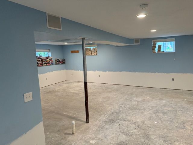 Blue and white painted unfinished basement with exposed concrete floor, support beams, and windows.