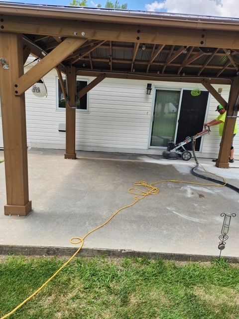 A worker grinding a concrete patio under a wooden pergola next to a white house with a sliding door.