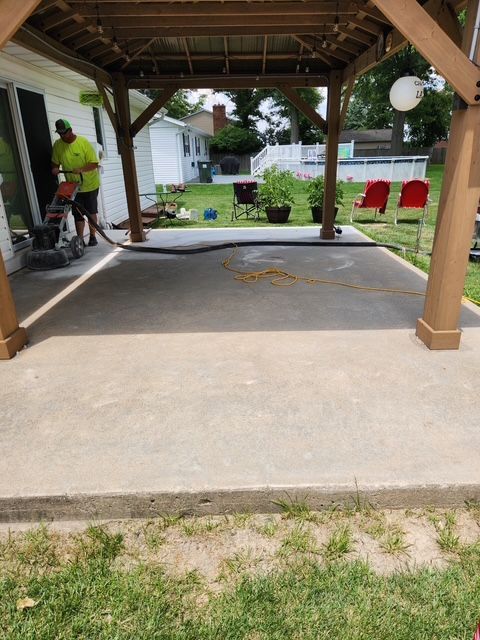 Man grinding a concrete patio under a wooden gazebo.  Green lawn, red chairs in background.