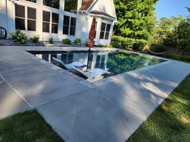 Pool with concrete surround, in a backyard. House in background, umbrella and lounge chairs in the water.