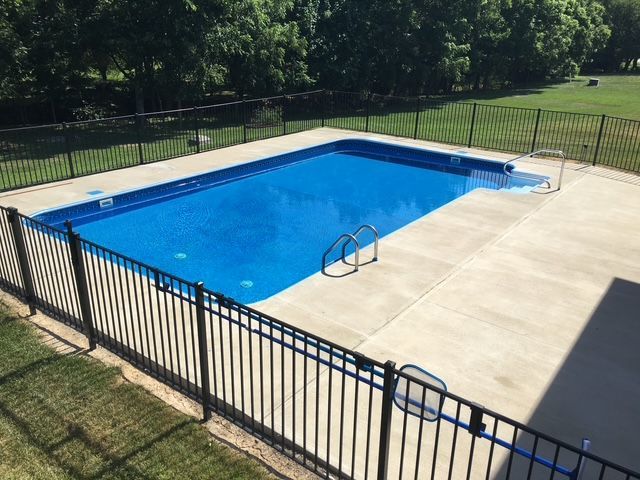 Rectangular in-ground swimming pool with blue water, concrete deck, and black metal fence in a yard with green grass and trees.