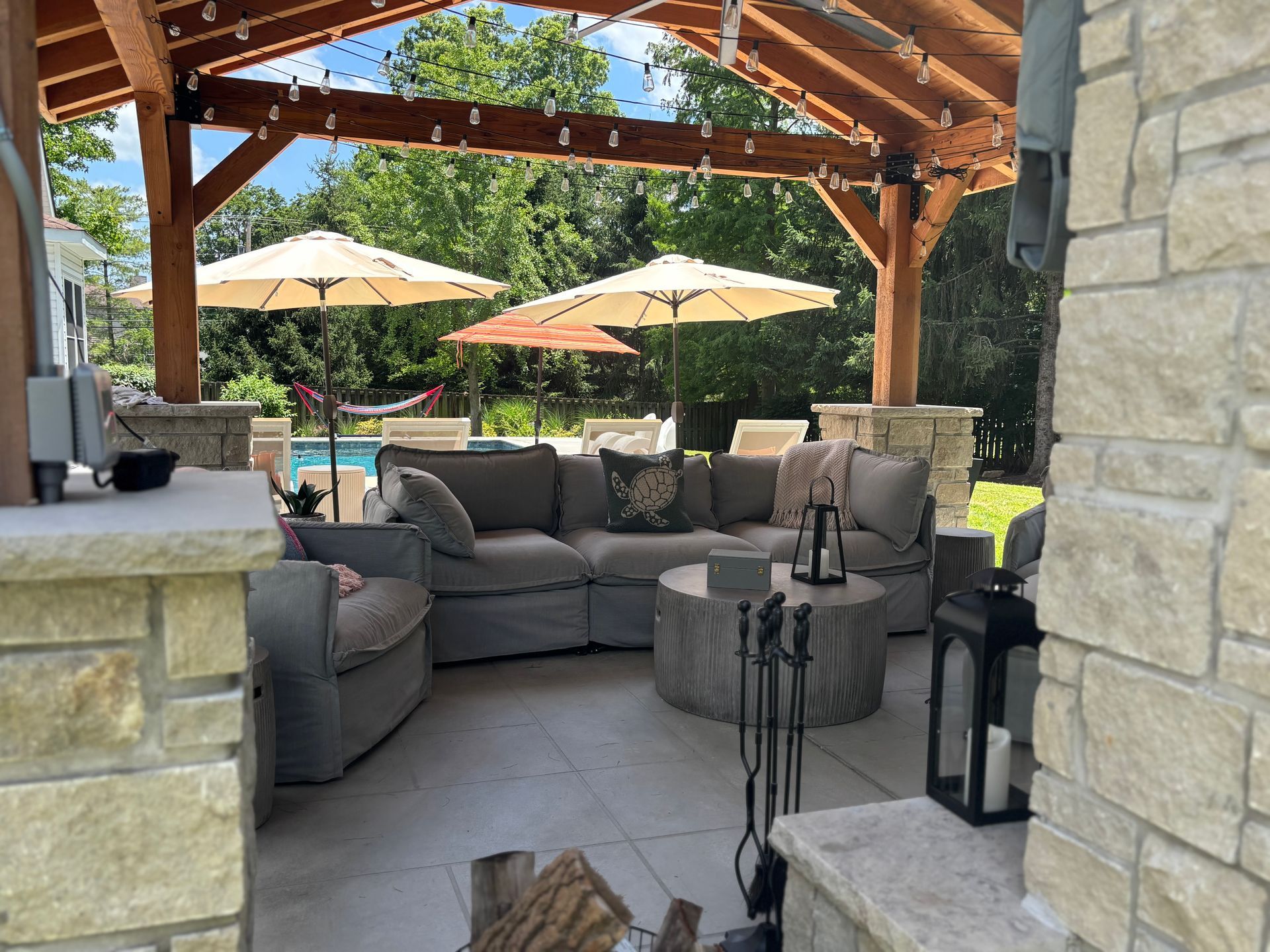 Patio with couches, umbrella-covered pool, stone facade, and overhead wooden beam structure.