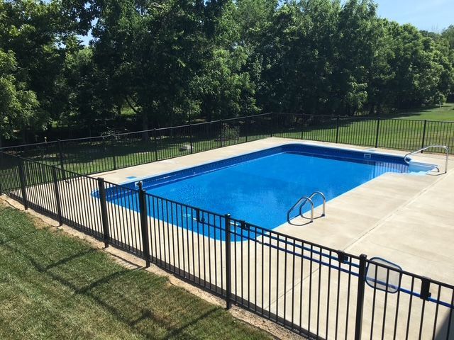 Rectangular in-ground pool surrounded by black fence and concrete. Green grass and trees are in the background.