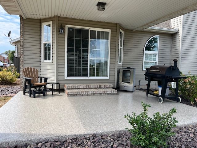 Patio with sliding glass door, grill, and chair on a concrete surface, beside a house with tan siding.