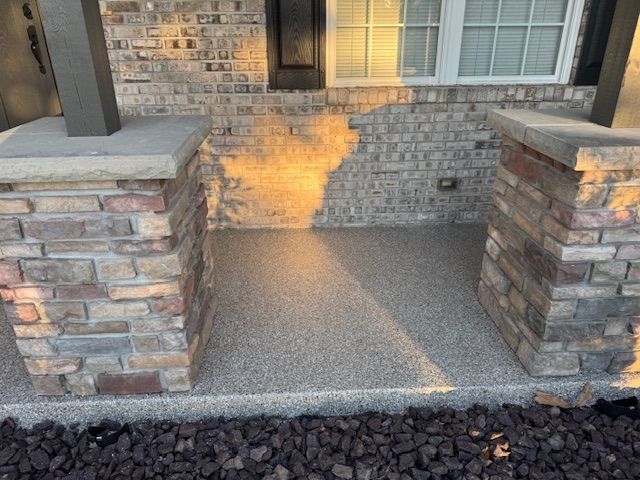 Stone pillars frame a small covered porch with aggregate flooring; brick wall and window in background.