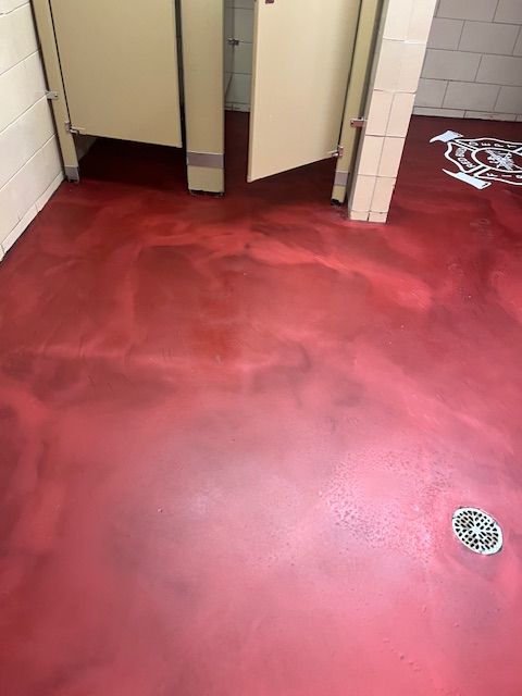 Red floor in a public restroom with stall doors open. There is a drain and a logo on the floor.