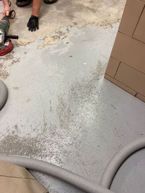 Grinding concrete floor. Worker wears gloves, angle grinder and vacuum hose visible. Gray concrete, beige debris.