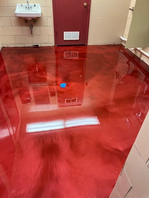 Red, glossy epoxy floor in a room with a red door, white sink, and beige walls, reflecting lights.