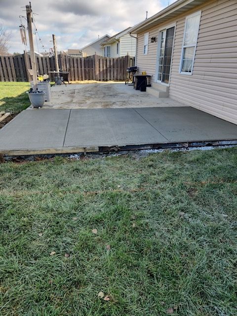 Newly poured concrete patio next to a light-colored house. Green grass in the foreground, wooden fence in background.