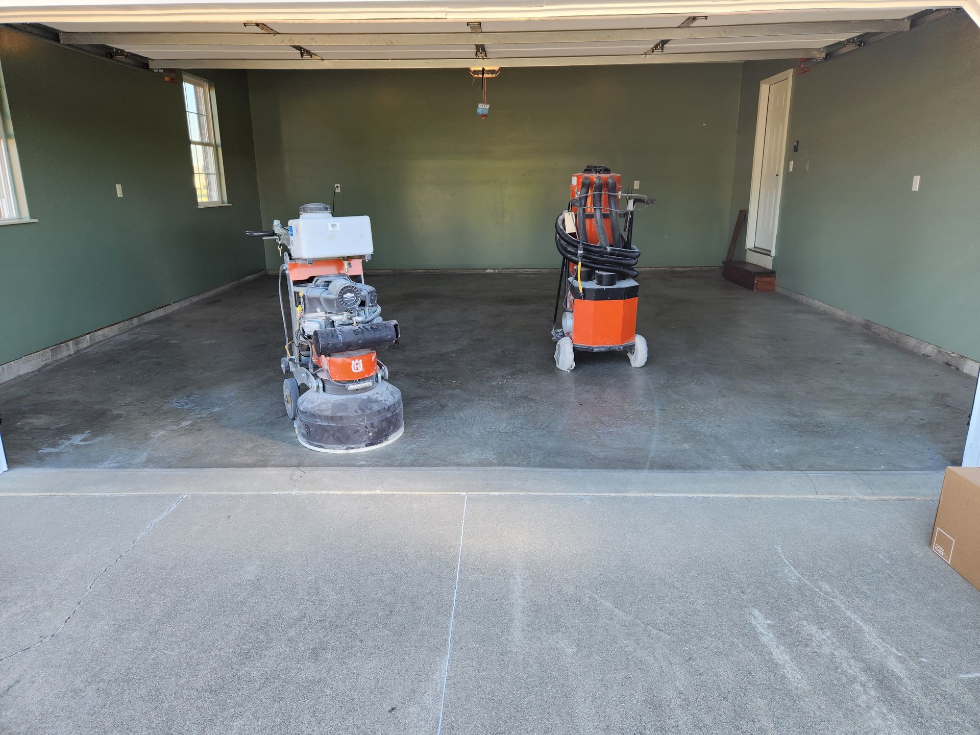 Two concrete grinders inside a garage. The floor is freshly ground, walls are olive green.
