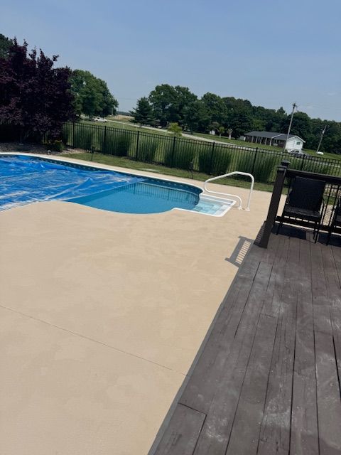 Pool with a blue cover, tan concrete deck, and wooden deck. Surrounded by a black fence and greenery.