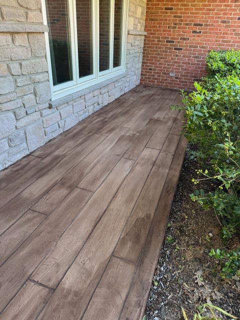 Concrete walkway stamped to resemble wood planks, next to stone and brick walls with green shrubs.