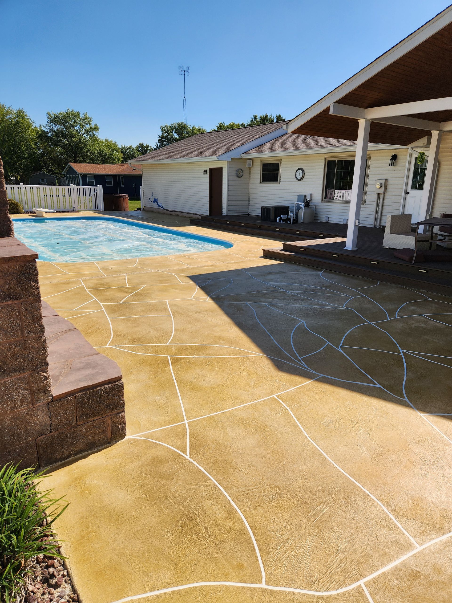 Backyard with a yellow stained concrete patio, pool, and white house. Sun is shining on a clear day.