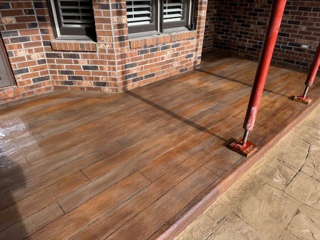 Concrete patio with a wood grain texture, stained in shades of brown and supported by red props, next to a brick building.