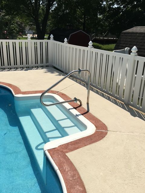 Pool steps with a metal handrail, surrounded by a white picket fence and concrete patio.