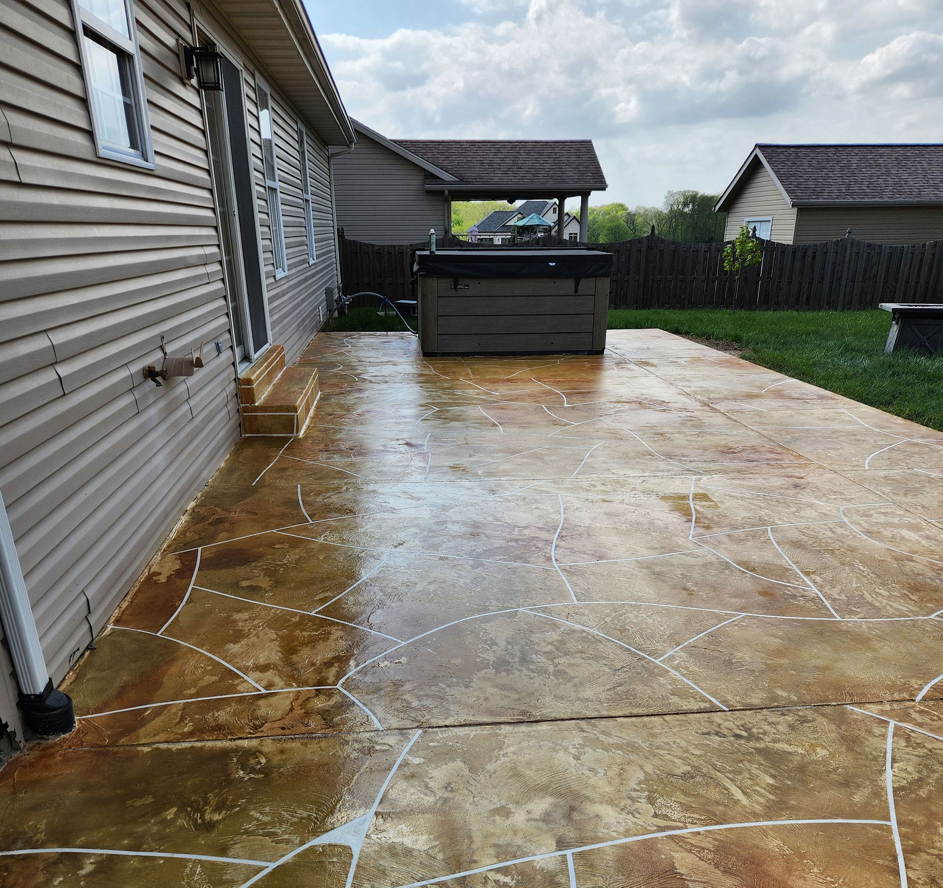Brown stained concrete patio with hot tub, next to a tan sided house and grass yard.