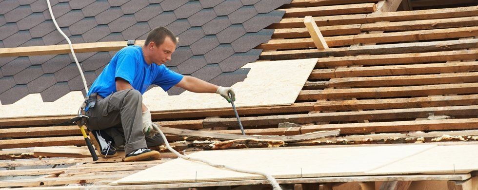 A roofer wearing safety gear is working on a roof, using a tool.