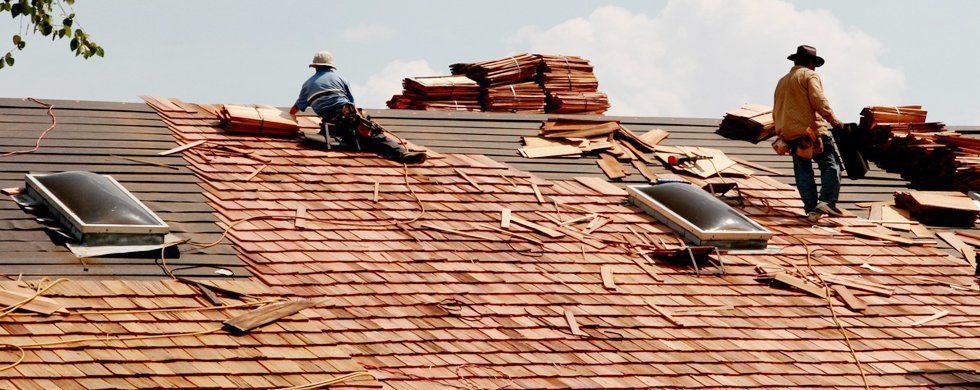 Roofers working on a roof, removing old shingles. Two skylights visible. Bright daylight.