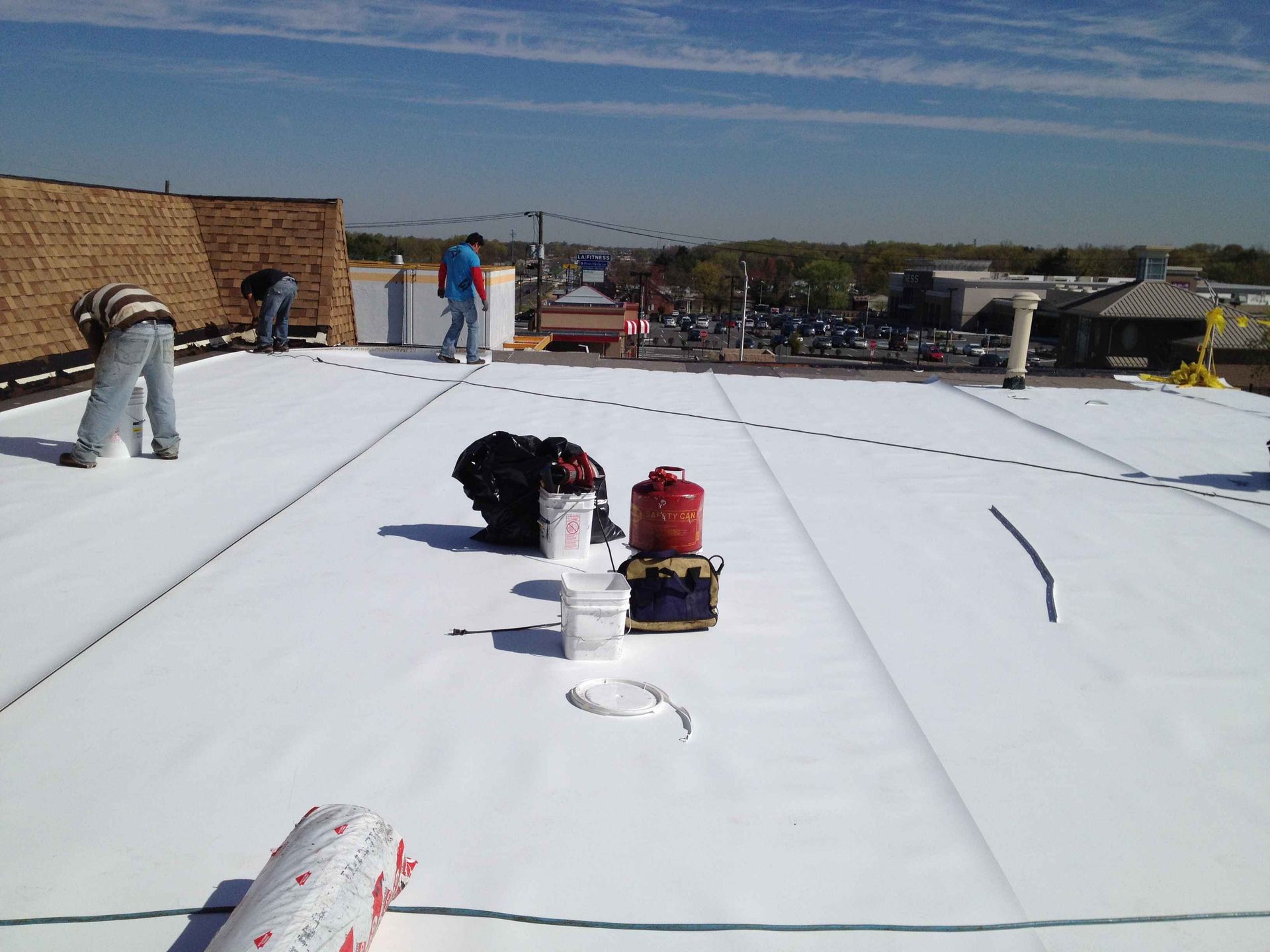 Workers installing white roofing material on a flat rooftop under a blue sky.