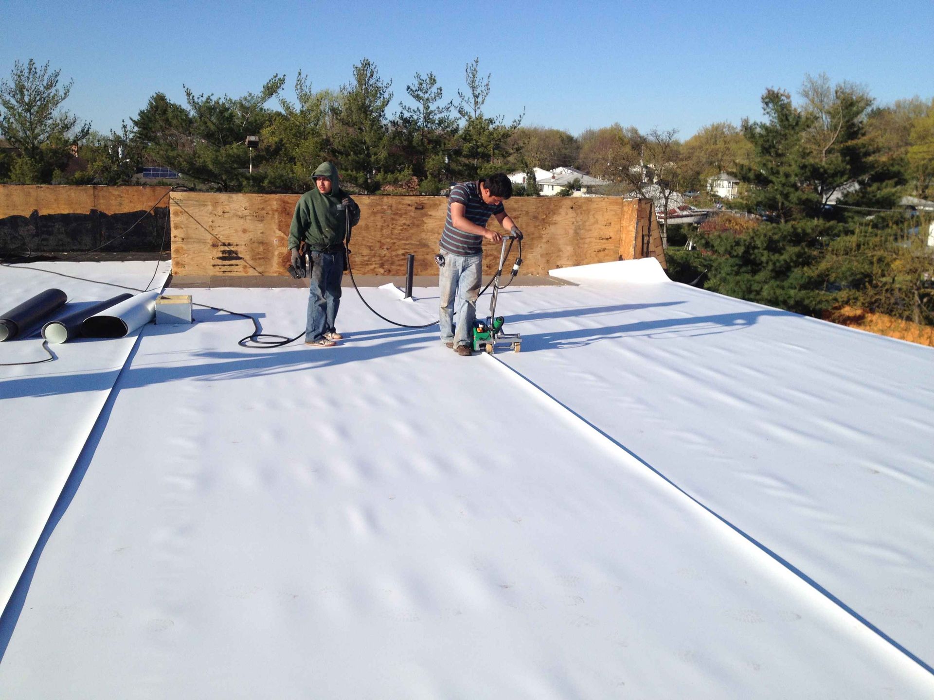 Two workers on a rooftop installing white roofing membrane.
