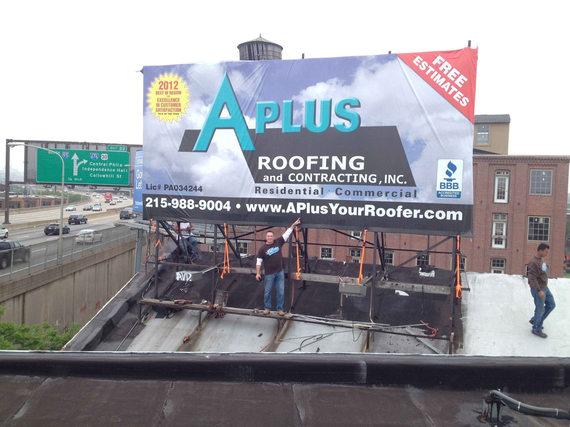 Billboard for A Plus Roofing on a roof; two people stand near it.