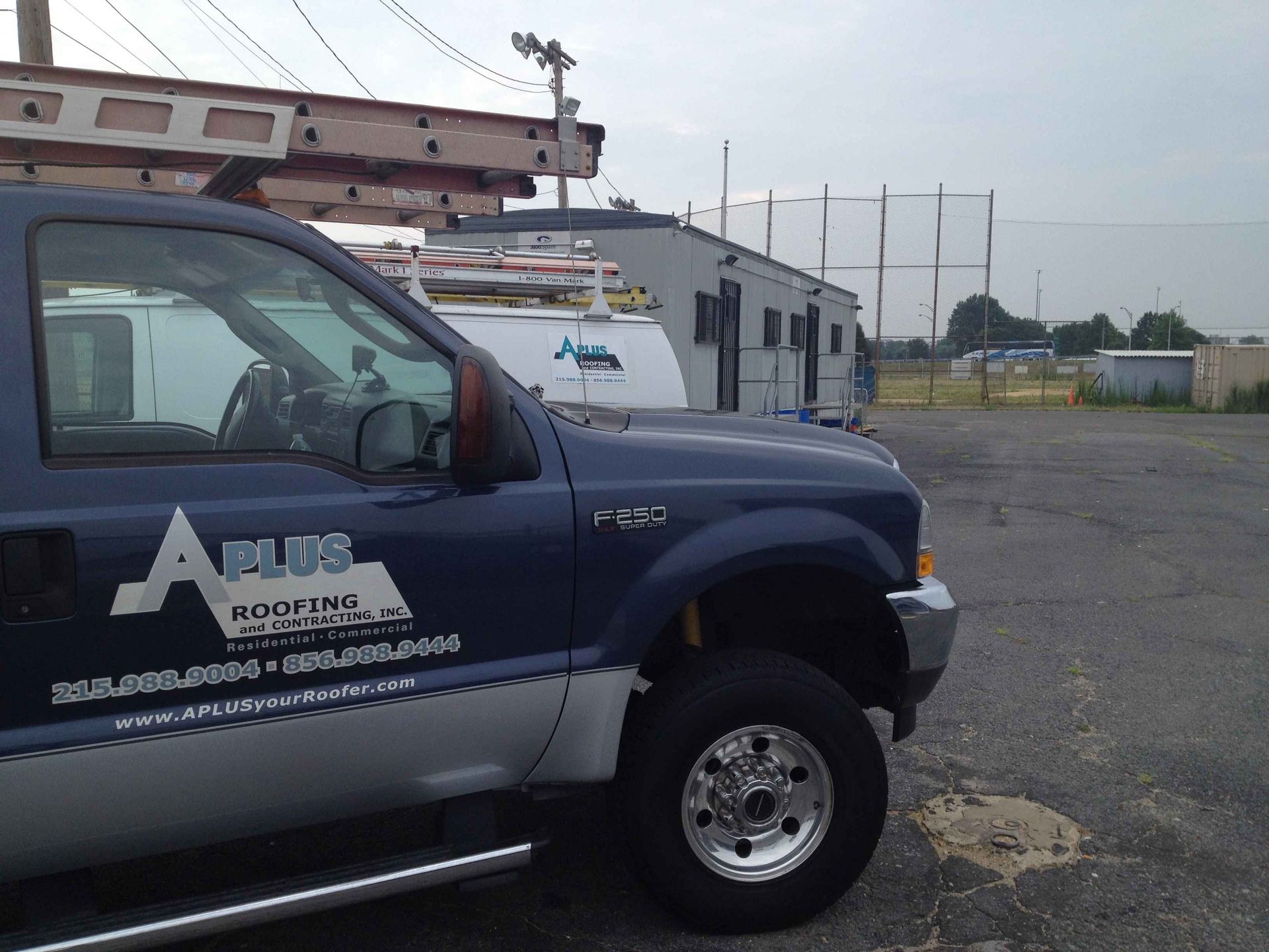Blue and silver roofing truck parked with a white work truck in the background. A field and cloudy sky are in the distance.