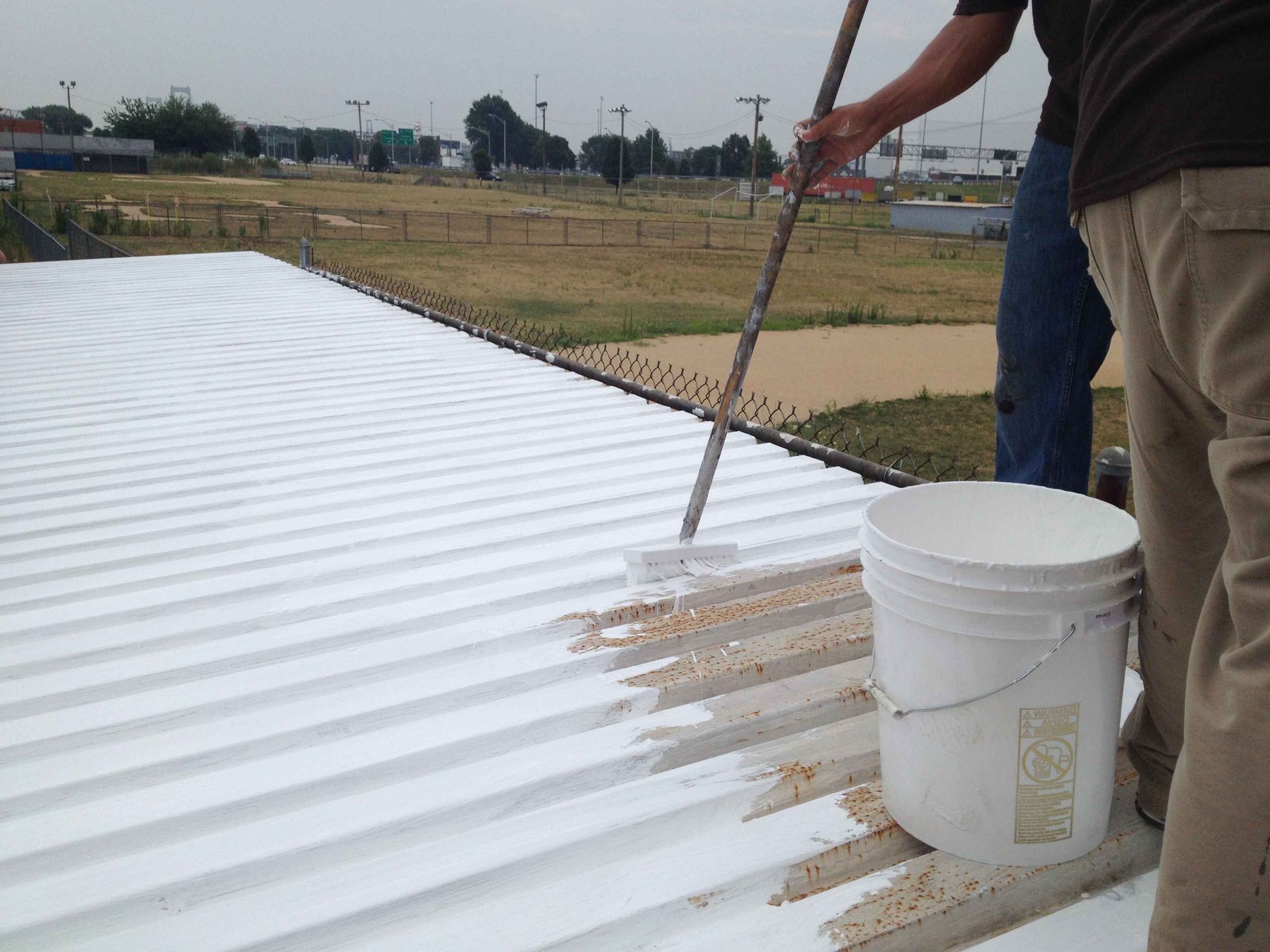 Person painting a white coating on a corrugated metal roof with a brush and bucket outdoors.