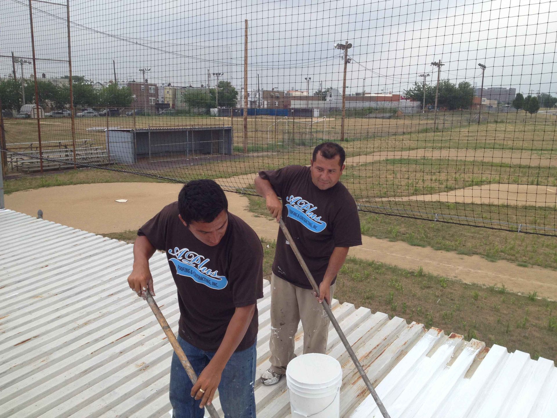 Two men in brown shirts sweeping a white corrugated roof, baseball field in the background.