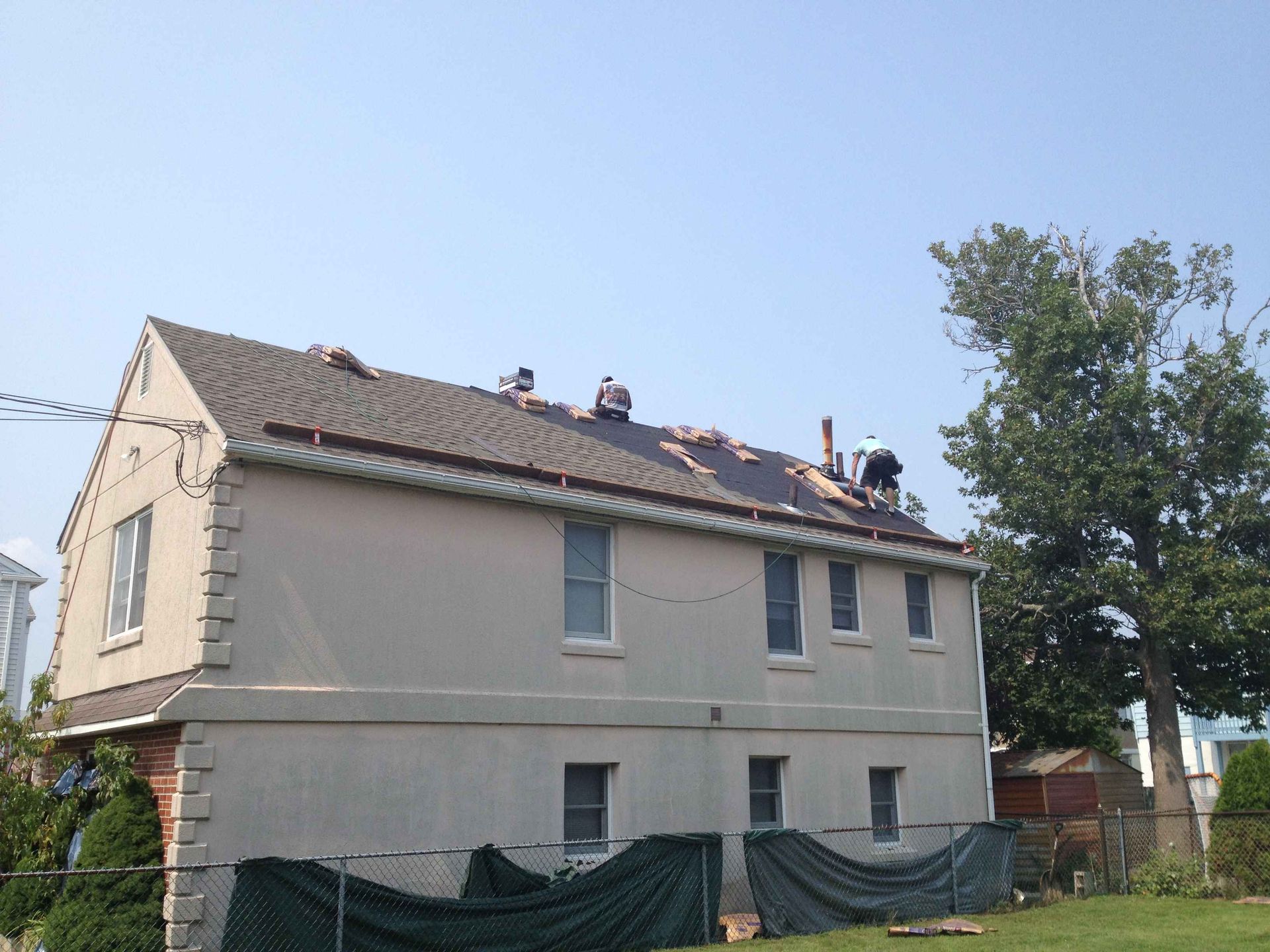 Workers replacing a roof on a two-story beige house under a clear, blue sky.