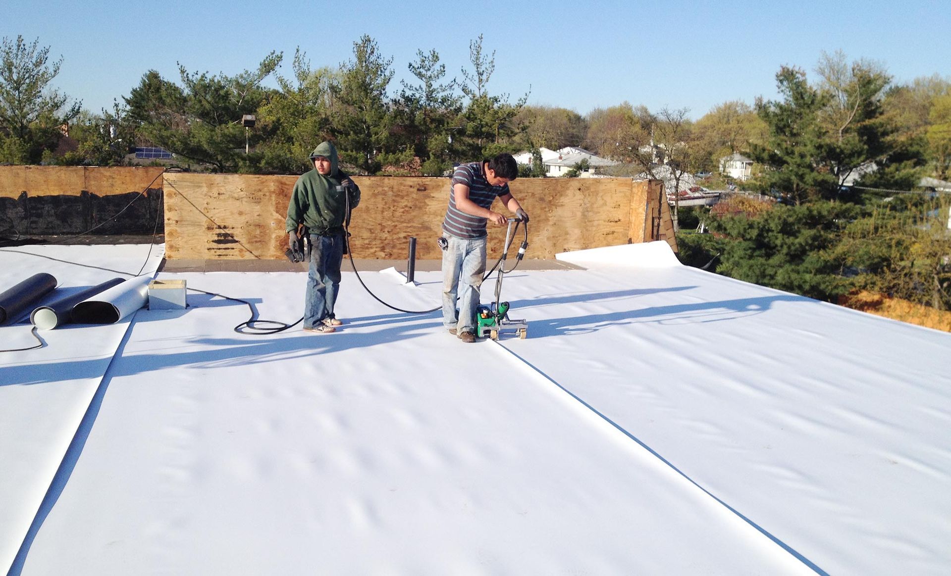 Two workers installing white roofing material on a flat roof; one operates a heat welder.