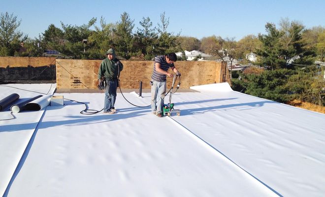 Two workers installing white roofing material on a flat roof; one operates a heat welder.