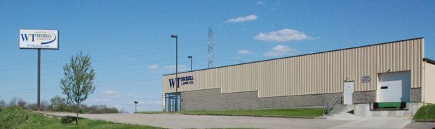 Industrial warehouse building on a sunny day, with a sign and a small door entrance beside a gravel lot.