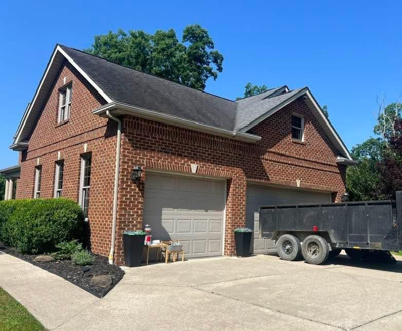 Two-story brick house with a two-car garage and a black trailer in the driveway on a sunny day.