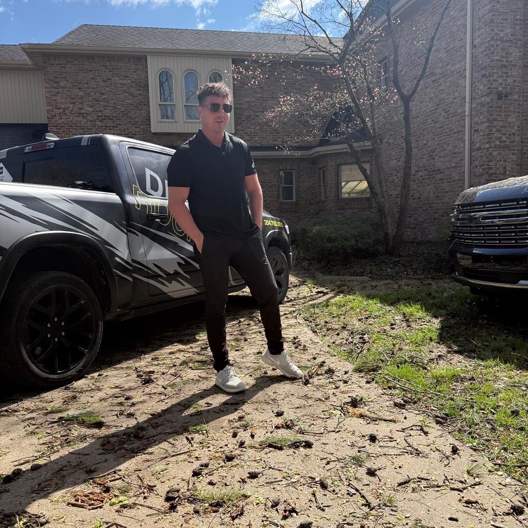 A man in a black polo shirt and pants stands between two vehicles in front of a brick house on a sunny day.