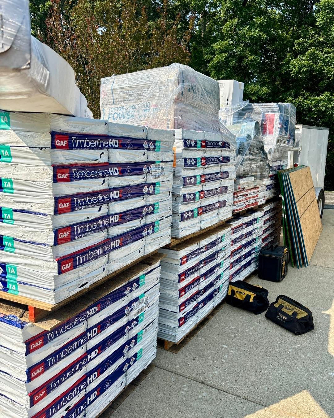 Stacks of roofing materials, including shingles, are arranged on pallets outdoors, with tool bags nearby.