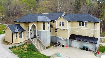 A two-story beige house with a dark roof, gray stone accents, and a driveway. The house features a unique tower and attached garage.