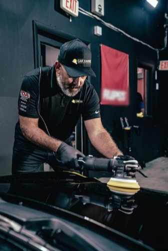 Man polishes car with a buffer in a garage; wearing a black shirt and cap.