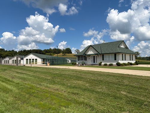 Wide grassy field with a white farmhouse and outbuildings under a blue sky with clouds