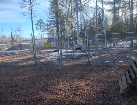 A metal fence surrounds a dirt field with trees in the background.