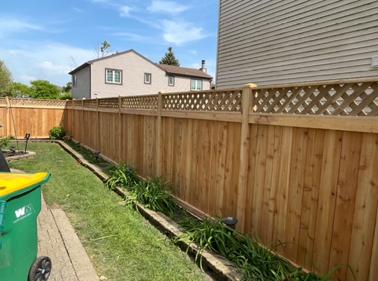 A wooden fence in a backyard with a green trash can in front of it.