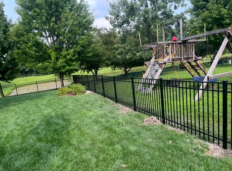 Black metal fence bordering a grassy backyard with a swing set on a slight hill, with trees and a fence in the background.