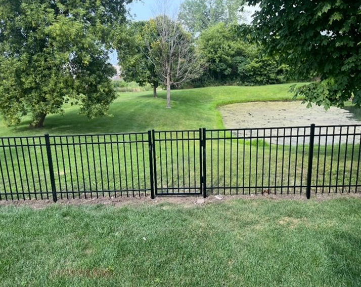 Black metal fence with a gate, surrounding a grassy area with a pond and trees in the background.