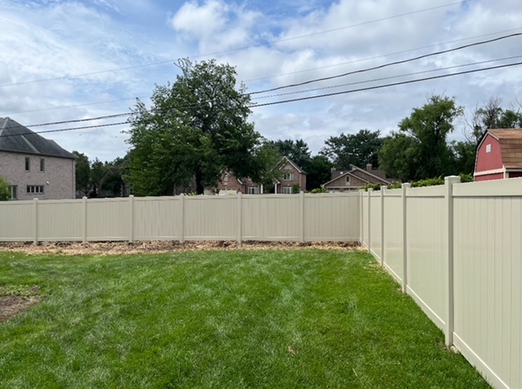 A beige vinyl fence encloses a grassy backyard with houses and trees visible beyond. The sky is partly cloudy.