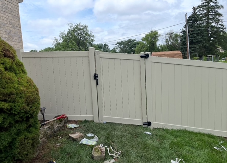 A beige vinyl fence with a gate in a backyard on a cloudy day. A green bush is on the left, and grass in the foreground.